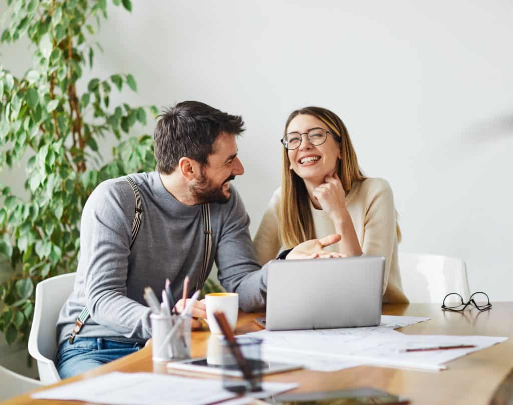 A happy couple sits at a table, engaged in a lively conversation while working on a laptop. The table is scattered with papers, a coffee mug, and pens, indicating they are working on a project together. Both are smiling and appear to be enjoying the moment.