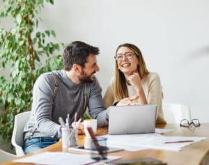 A happy couple sits at a table, engaged in a lively conversation while working on a laptop. The table is scattered with papers, a coffee mug, and pens, indicating they are working on a project together. Both are smiling and appear to be enjoying the moment.