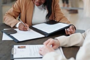 two people sitting at a table, reviewing and signing documents. One person, wearing a light brown jacket, is holding a pen and signing a document that is in a black folder. The other person, whose hands are visible, is also looking over documents on the table.