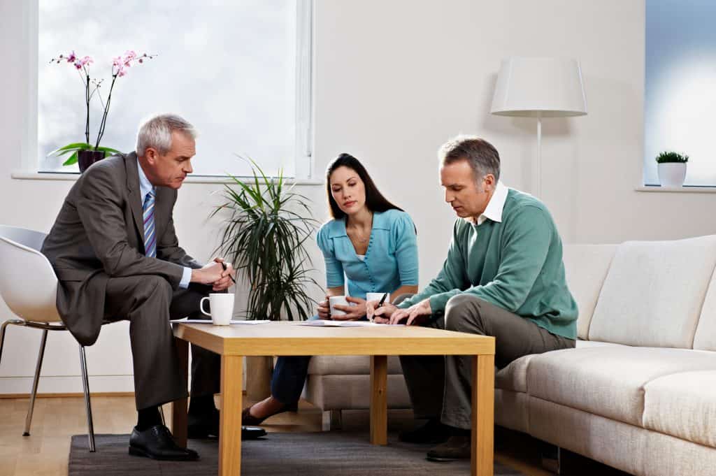 The image shows a middle-aged couple sitting on a couch in a modern, well-lit living room, discussing documents with an older, professionally dressed man. The man appears to be advising the couple, possibly in a legal or financial capacity, as they review paperwork together.