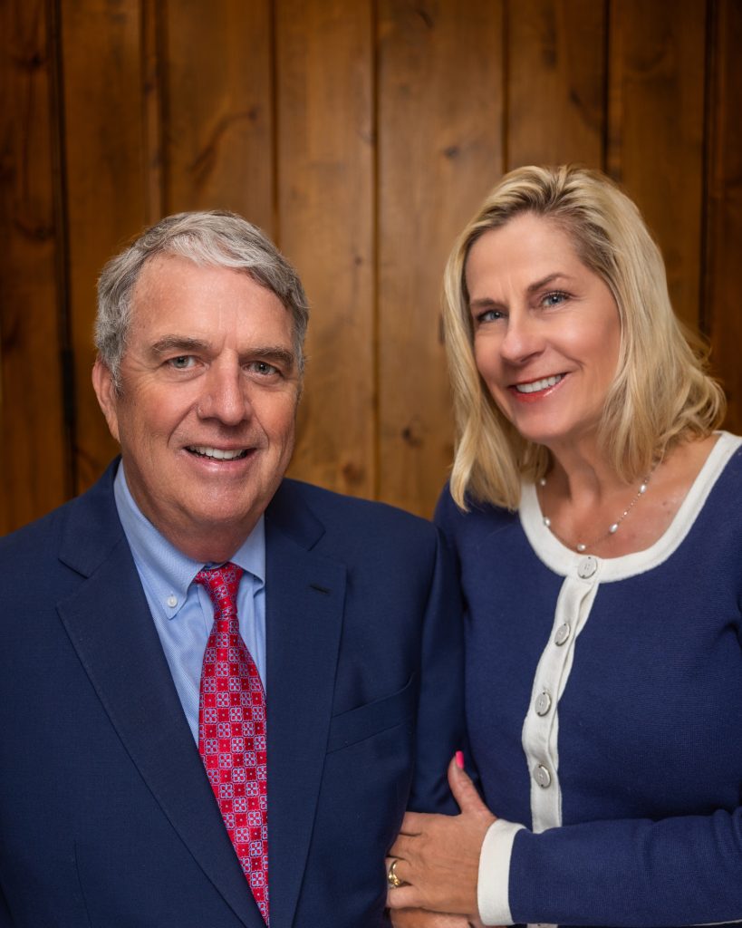 A portrait of Matt Dana and his wife smiling together against a wood-paneled background. Matt, on the left, has gray hair and wears a navy blue suit with a red patterned tie. His wife, on the right, has blonde hair and wears a blue cardigan with white trim and buttons, resting her hand gently on his arm.