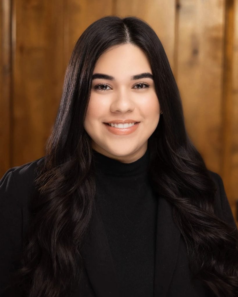 Aylza Ruiz, a paralegal at Dana Whiting Law, poses with long dark hair, smiling, wooden background.