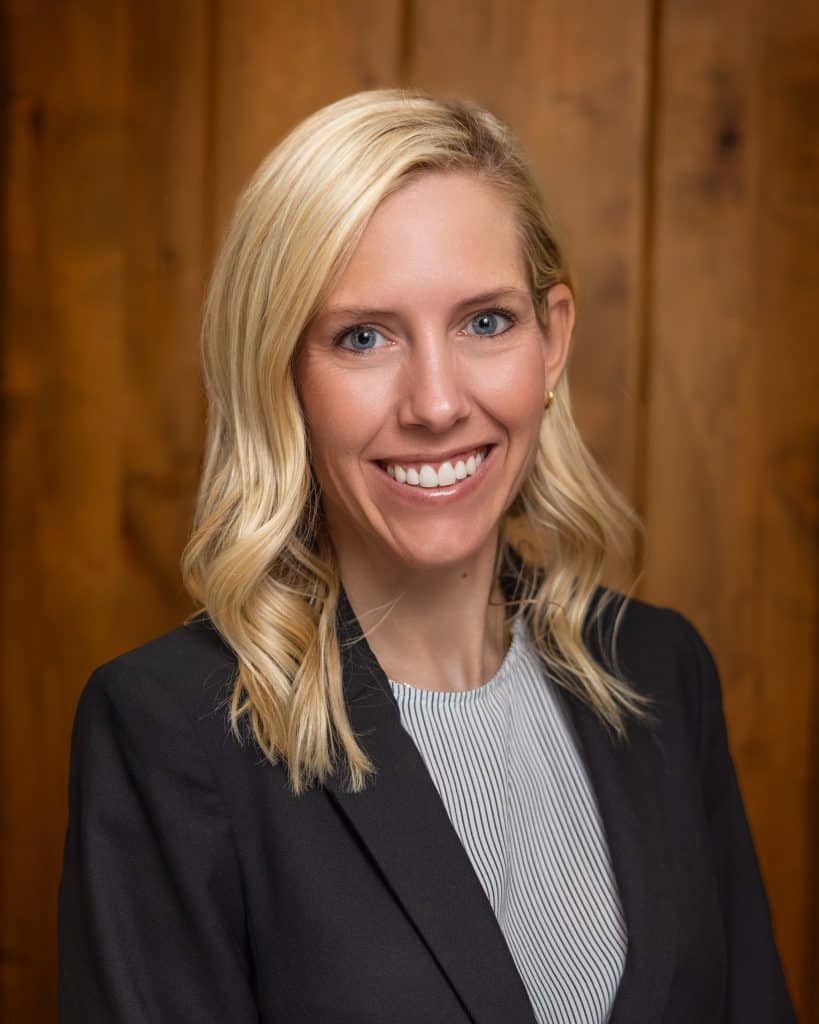 A portrait of Hallie Tessendorf smiling warmly at the camera. She has shoulder-length, wavy blonde hair and blue eyes, and is wearing a black blazer over a light-colored, finely striped shirt, standing against a wood-paneled background.
