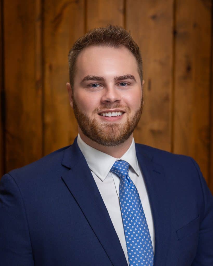 Michael B. Rich, Legal professional portrait of a young man in a navy suit and blue tie, smiling confidently against a wooden background.