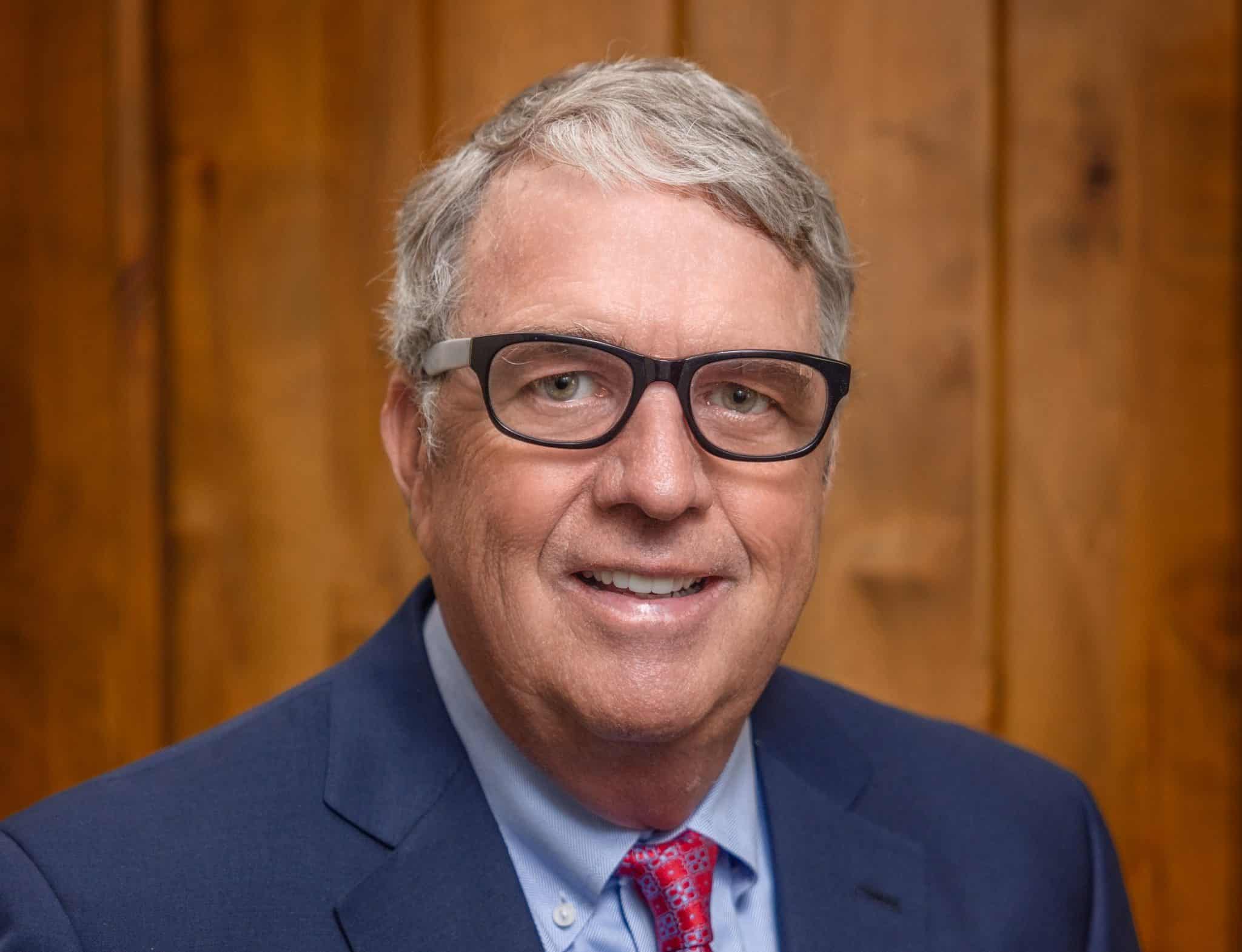 A close-up portrait of Matt Dana wearing black-framed glasses, a navy blue suit jacket, a light blue button-down shirt, and a red patterned tie, standing against a background of vertical wood paneling.